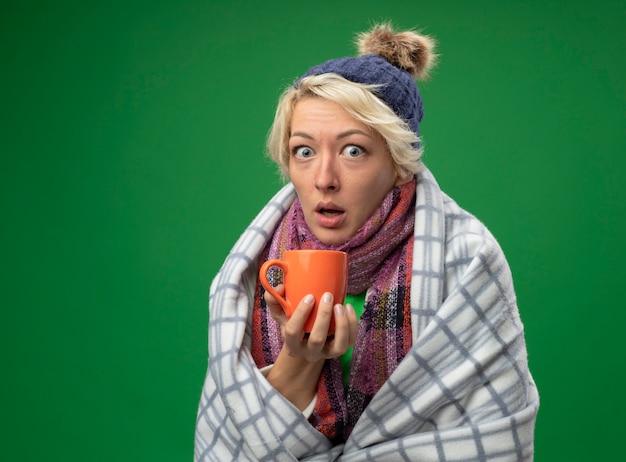 Donna malata malsana con i capelli corti in sciarpa calda e cappello sensazione di malessere avvolto in una coperta che tiene tazza di tè caldo guardando la telecamera essendo sorpreso in piedi su sfondo verde