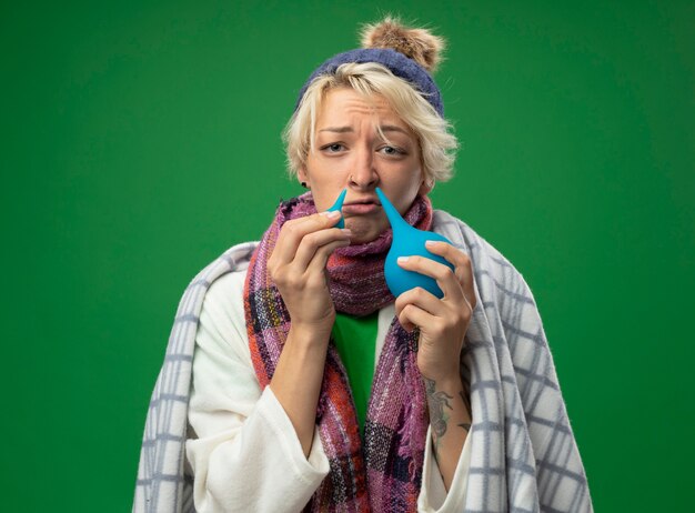 Sick unhealthy woman with short hair in warm scarf and hat feeling unwell wrapped in blanket holding enemas near her noseover green wall