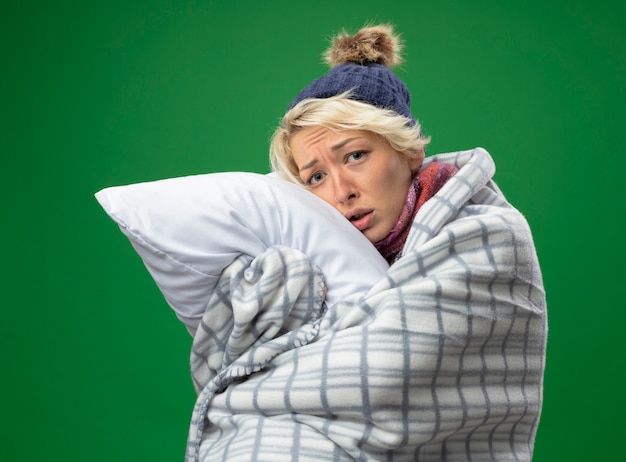 Sick unhealthy woman with short hair in warm scarf and hat feeling unwell wrapped in blanket holding pillow looking at camera with sad expression standing over purple background