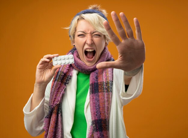 Sick unhealthy woman with short hair in warm scarf and hat holding blister with pills making stop gesture with hand shouting standing over orange background