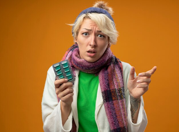 Sick unhealthy woman with short hair in warm scarf and hat holding blister with pills pointign with index finger to the side looking confused standing over orange wall
