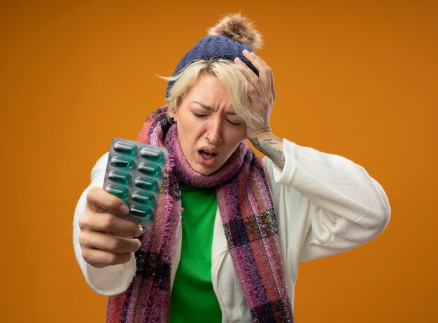 Sick unhealthy woman with short hair in warm scarf and hat holding blister with pills touching her head looking confused standing over orange wall