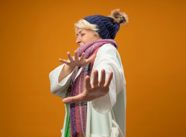 Foto gratuita donna malsana malata con capelli corti in sciarpa calda e cappello che fa gesto di difesa con le mani in piedi su sfondo arancione