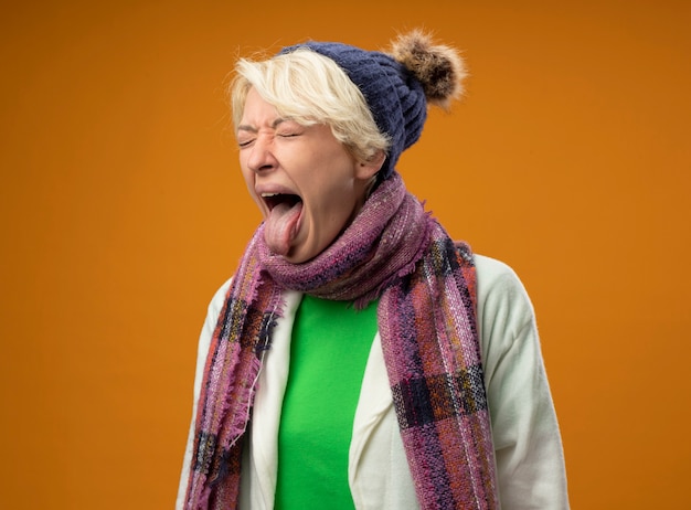 Sick unhealthy woman with short hair in warm scarf and hat sticking out togue feeling unwell standing over orange wall