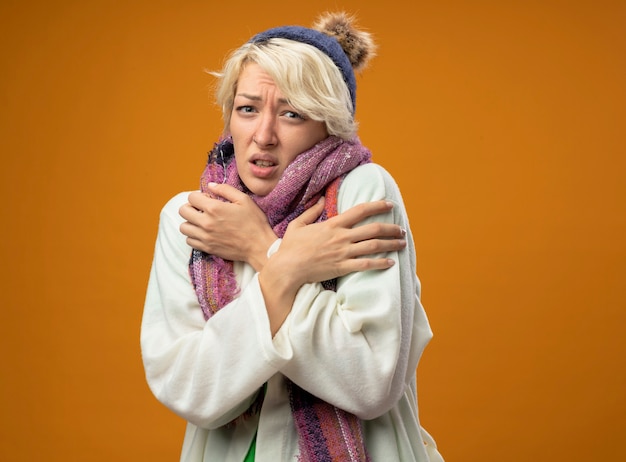 Sick unhealthy woman with short hair in warm scarf and hat suffering from fever feeling unwell standing over orange wall