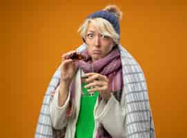 Free photo sick unhealthy woman with short hair in warm scarf and hat wrapped in blanket dripping medicine drops into a glass looking at camera confused standing over orange background