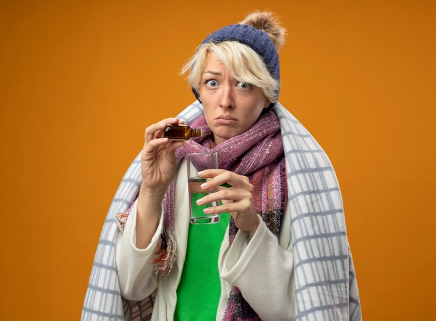 Sick unhealthy woman with short hair in warm scarf and hat wrapped in blanket dripping medicine drops into a glass looking at camera confused standing over orange background