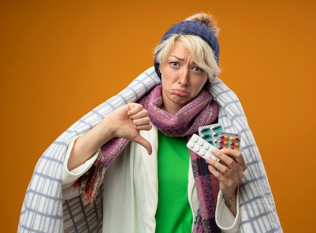 Sick unhealthy woman with short hair in warm scarf and hat wrapped in blanket holding pills looking at camera unhappy and sad showing thumbs down standing over orange background