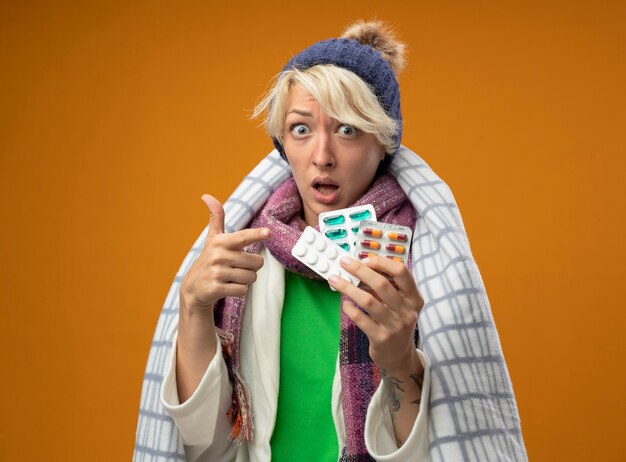 Больная нездоровая женщина с короткими волосами в теплом шарфе и шляпе, завернутой в одеяло, держит пуантин с таблетками, с пальцем на них обеспокоенная, стоя над оранжевой стеной