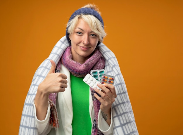 Sick unhealthy woman with short hair in warm scarf and hat wrapped in blanket holding pills smiling showing thumbs up feeling better standing over orange wall