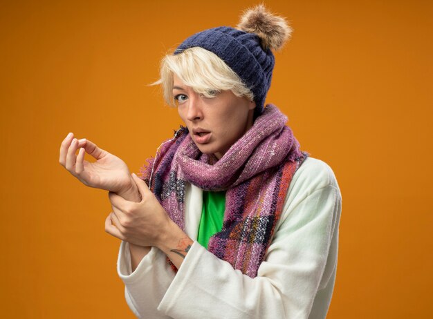 Sick unhealthy woman with short hair in warm scarf and hatlistening to her pulse looking worried standing over orange wall