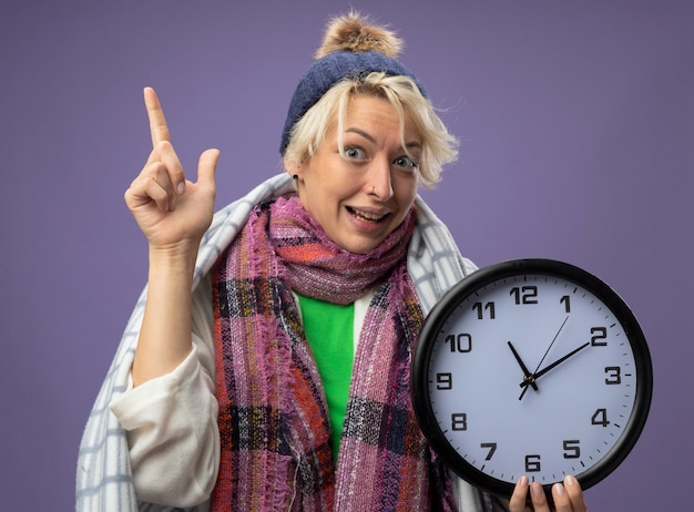 Foto gratuita donna malsana malata con capelli corti in sciarpa calda e cappello avvolto in una coperta che tiene l'orologio da parete che si sente meglio mostrando il dito indice che sorride sulla parete viola