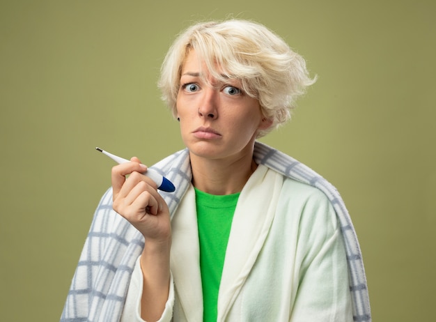 Free photo sick unhealthy woman with short hair wrapped in blanket feeling unwell holding thermometer looking at camera with sad expression suffering from flu standing over light background