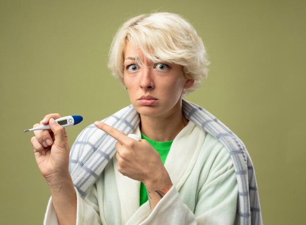 Sick unhealthy woman with short hair wrapped in blanket feeling unwell holding thermometer pointing with index finger at it with sad expression suffering from flu