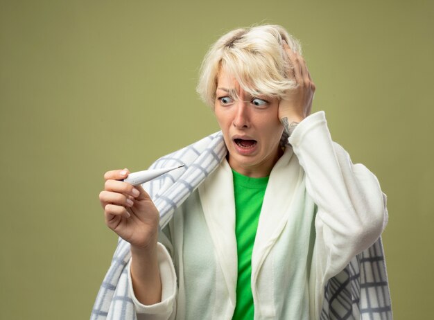 Foto gratuita malata malsana donna con i capelli corti avvolti in una coperta sensazione di malessere guardando il termometro essendo scioccato in piedi sopra la parete chiara