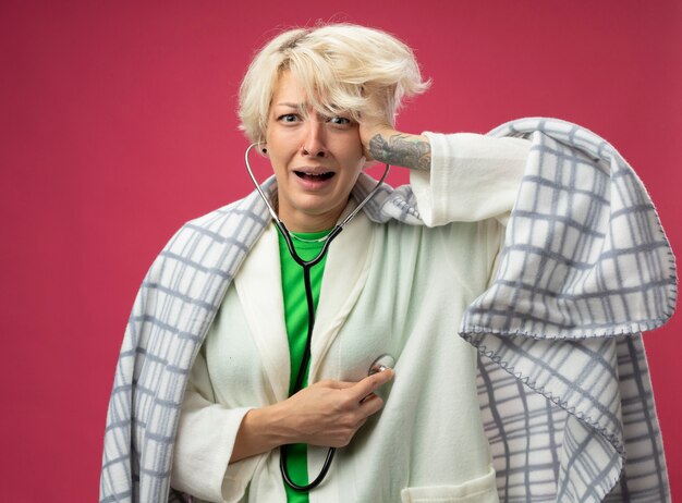 Sick unhealthy woman with short hair wrapped in blanket with stethoscope around her neck listening to her heartbeat feeling unwell stressed and nervous standing over pink background