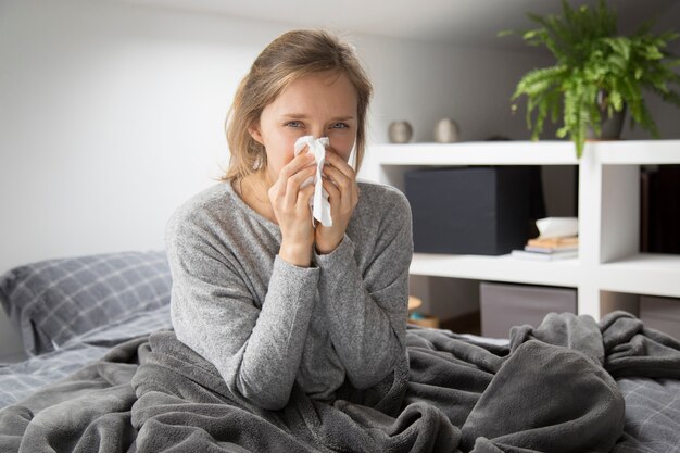 Sick woman in bed blowing nose with napkin, looking at camera