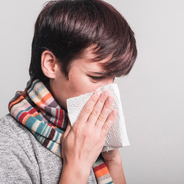 Sick woman blowing nose in tissue paper against gray background