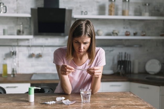 Sick woman checking temperature on thermometer with medicines on wooden desk
