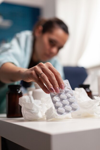 Sick woman having seasonal illness at home sitting in blanket on couch young adult taking medicine a...