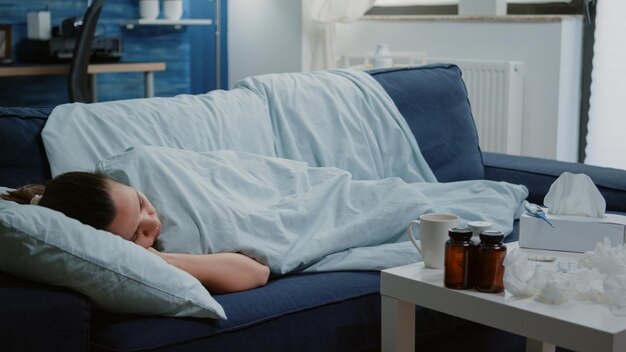 Больная женщина, лежащая на диване с одеялом, чувствует холод и симптомы вируса. Взрослый с сезонным гриппом спит, принимая лекарства на столе. Усталый больной человек с ознобом и дрожью