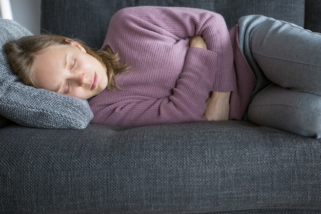 Больная женщина лежит на сером диване у себя дома, согнув ноги