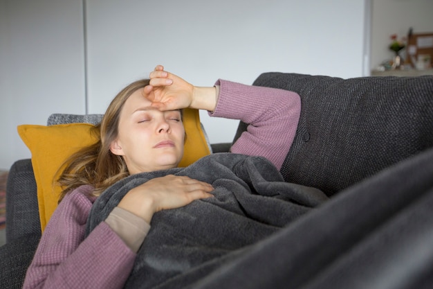 Больная женщина лежит на сером диване у себя дома, держась за руки на груди