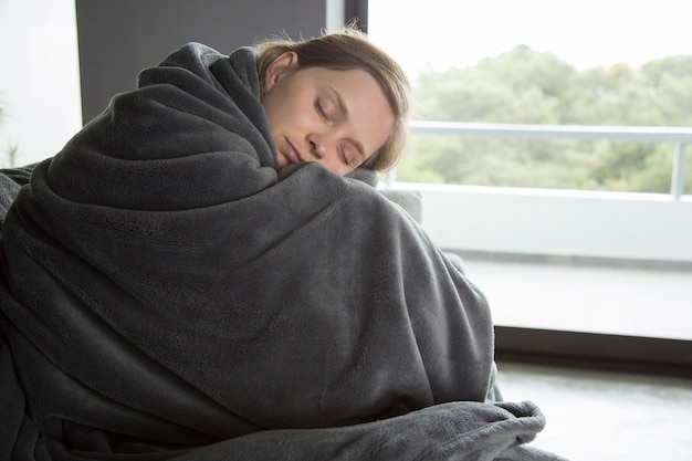 Sick woman sitting on sofa with closed eyes, embracing knees