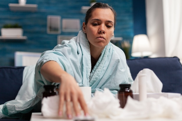 Sick woman taking medicine for seasonal virus wrapped in blanket holding pills caucasian young perso...