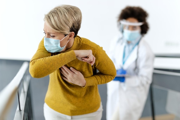 Free photo sick woman with face mask coughing into elbow at medical clinic