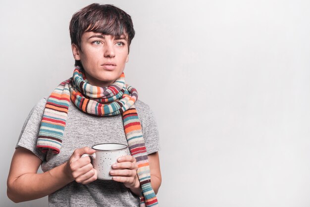 Sick woman with multicolored scarf around her neck holding mug against gray background
