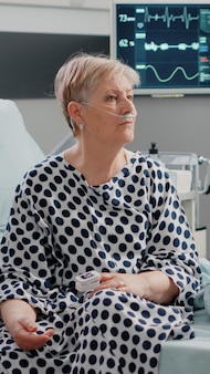 ヘルスケア​と​回復​の​ために​ベッド​に​座っている​鼻​酸素​チューブ​と​点滴​バッグ​を​持った​病気​の​女性​。​パルスオキシメータ​と​心拍​数​モニター​を​使用し​ながら​治療​を​待っている​退職した​患者