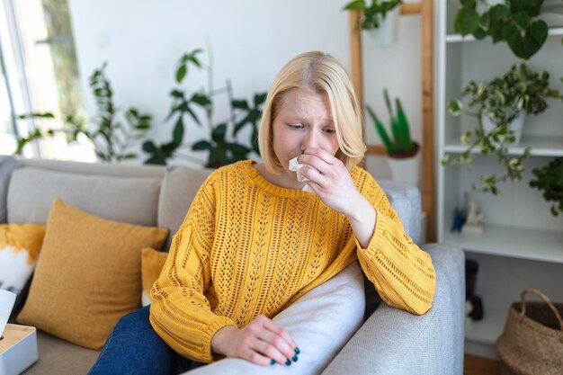 Sick WomanFluWoman Caught Cold Sneezing into Tissue Headache Virus Medicines Young Woman Infected With covid 19 Blowing Her Nose In Handkerchief Sick woman with a headache sitting on a sofa