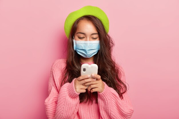 Sick young brunette woman with dark hair, focused in cellphone device, wears medical mask, has problems with health