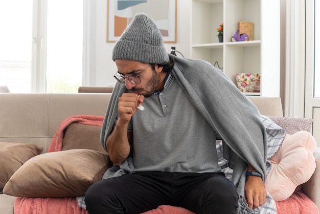 Free photo sick young caucasian man in optical glasses wearing winter hat coughing keeping fist close to mouth sitting on couch at living room