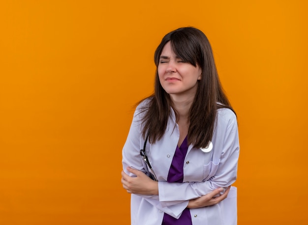 Sick young female doctor in medical robe with stethoscope holds belly with hands on isolated orange background with copy space