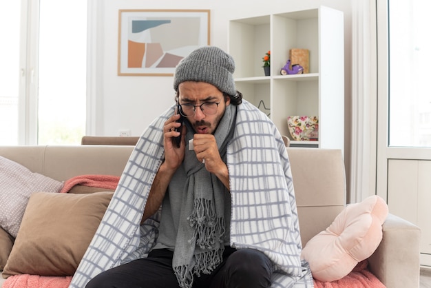 sick young man in optical glasses wrapped in plaid with scarf around his neck wearing winter hat coughing talking on phone sitting on couch at living room