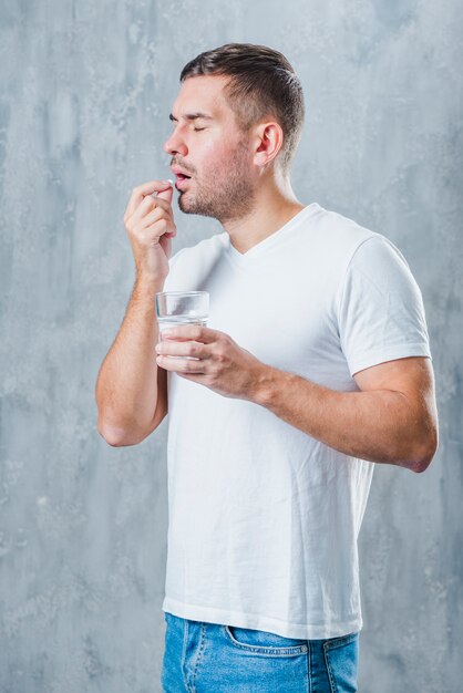 Sick young man standing against gray backdrop holding water glass in hand taking pill