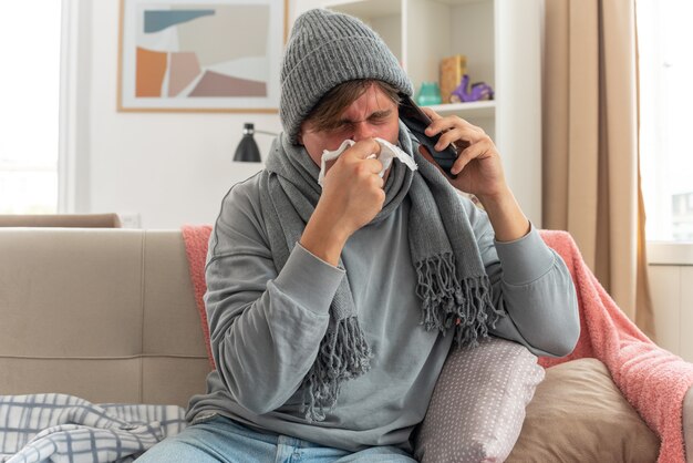 冬の帽子をかぶって首にスカーフを持っている病気の若い男は彼の鼻を拭き、リビングルームのソファに座って電話で話している