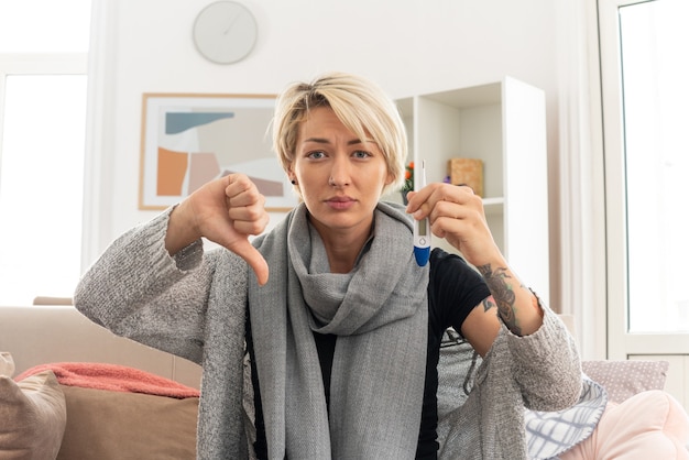 温度計を保持し、リビングルームのソファに座って親指を下に彼女の首の周りにスカーフを持つ病気の若いスラブ女性