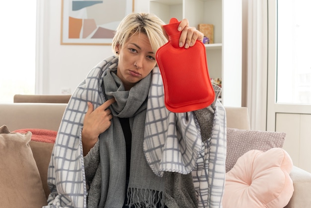 Free photo sick young slavic woman with scarf around her neck wrapped in plaid holding hot water bottle and pointing at herself sitting on couch at living room