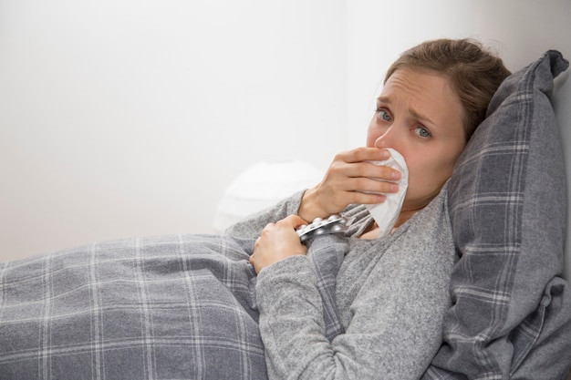 sick young woman in bed sneezing, holding pills in hand