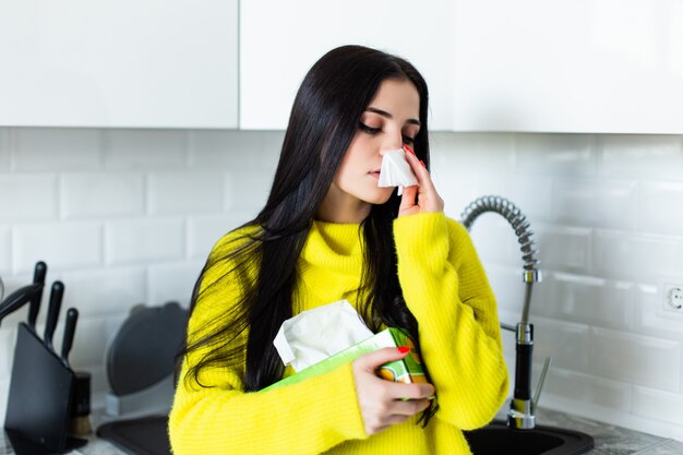 Sick young woman blows her nose in the kitchen.