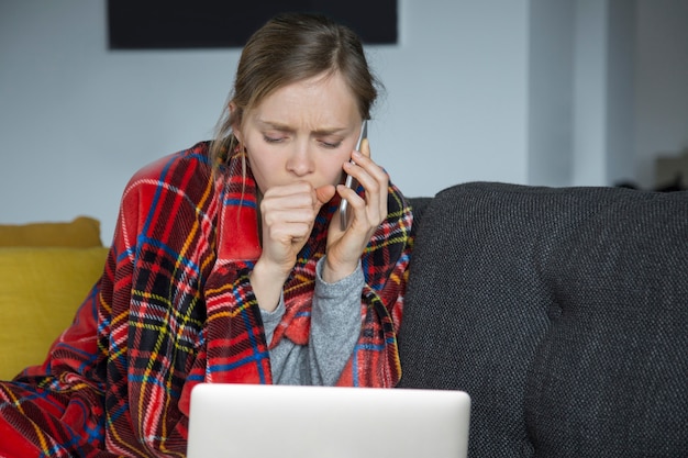 Sick young woman coughing, sitting at home, working on laptop