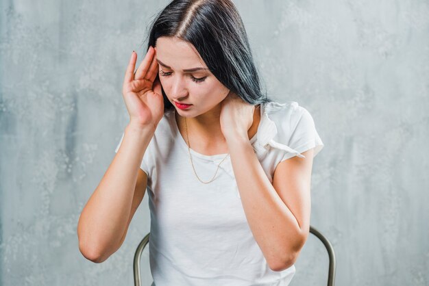 Sick young woman sitting against gray backdrop suffering from headache