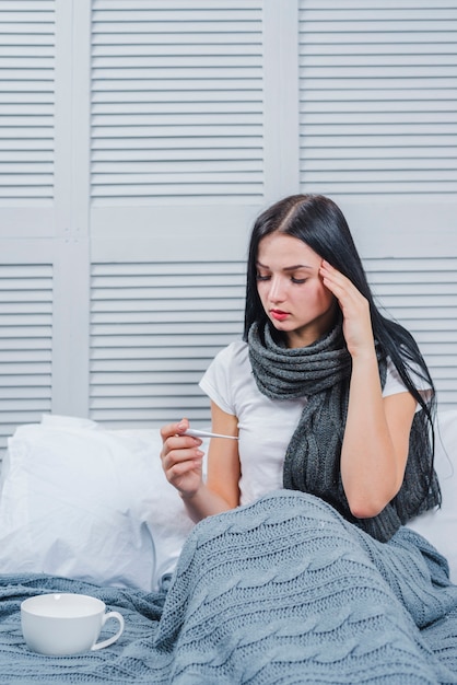 Sick young woman sitting on bed looking at thermometer