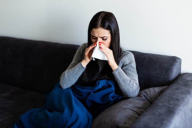 Sick young woman sitting on sofa blowing her nose at home in the sitting room