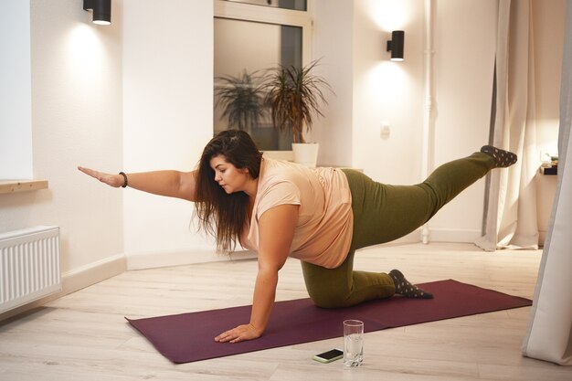 Sid view of overweight obese young woman wearing t-shirt and leggings doing physical training on mat to strengthen legs, arms, abs and spine. Weght loss, fitness, sports and active lifestyle concept