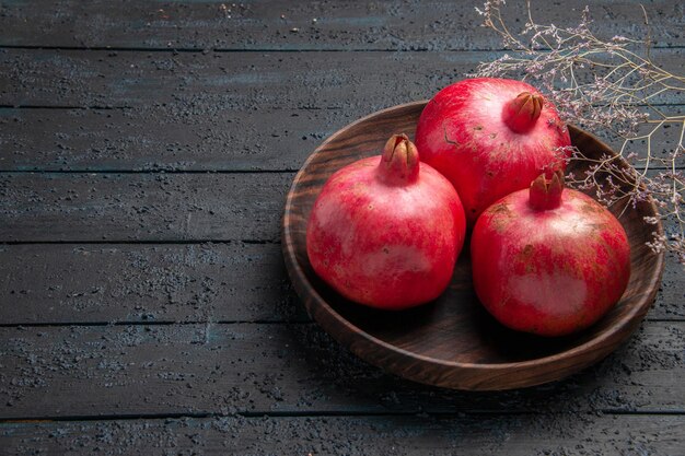 Side close-up view bowl of pomegranates three red pomegranates in bowl next to the branches on grey table