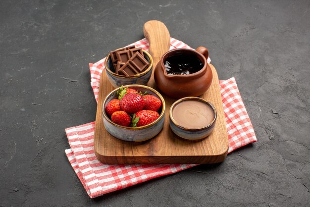 Side close-up view bowls on board berries and chocolate cream in bowls on the wooden cutting board on the pink-white checkered tablecloth on the dark table